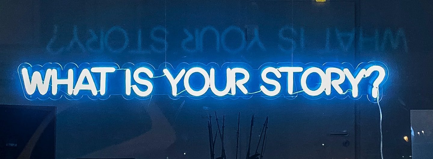 Foto versinnbildlicht mit der Neonschrift "What is your story?" die Stimmen zu Housing First Köln